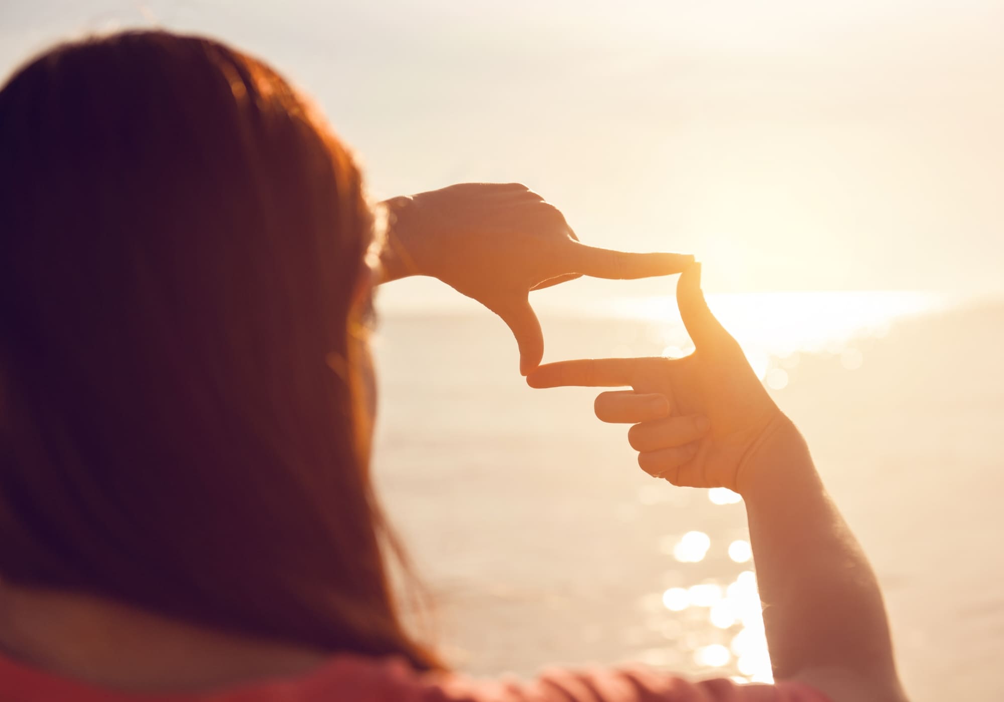 Person am Meer, die mit den Händen ein Rechteck formt und in die untergehende Sonne blickt. Warme Lichtstrahlen spiegeln sich im Wasser und erzeugen eine inspirierende, verträumte Atmosphäre.