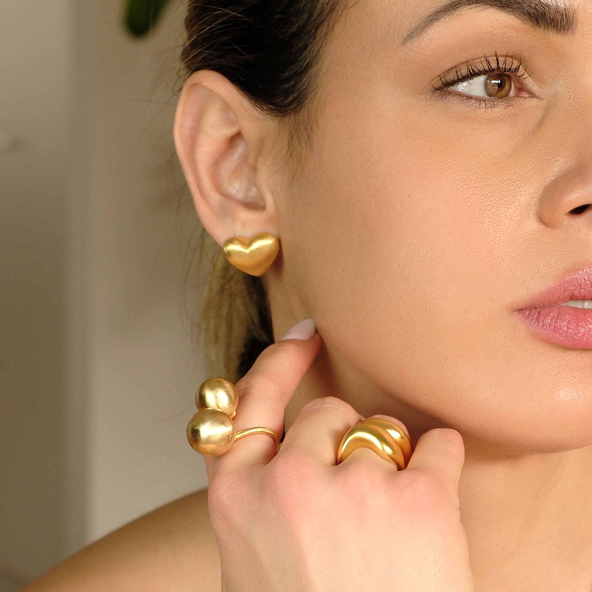 Model trägt die Cosytas Lumina Gold Heart Ohrringe. Zeitlose, herzförmige Ohrstecker mit sanfter, gebürsteter Oberfläche. Kombiniert mit passenden Statement-Ringen für einen eleganten, modernen Look.