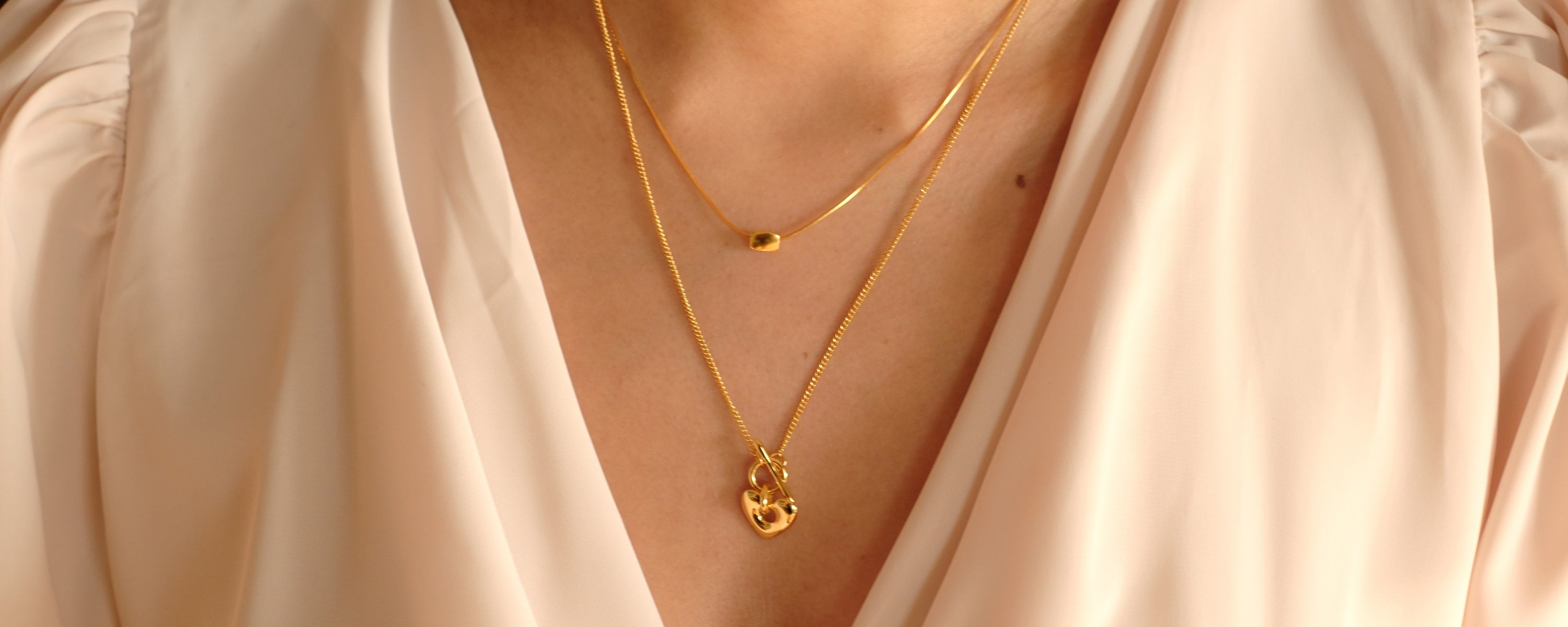 Frau trägt zwei goldene Halsketten in Layering-Optik über einem eleganten cremefarbenen Oberteil. Die Ketten haben feine Anhänger und verleihen dem Look eine edle Note.