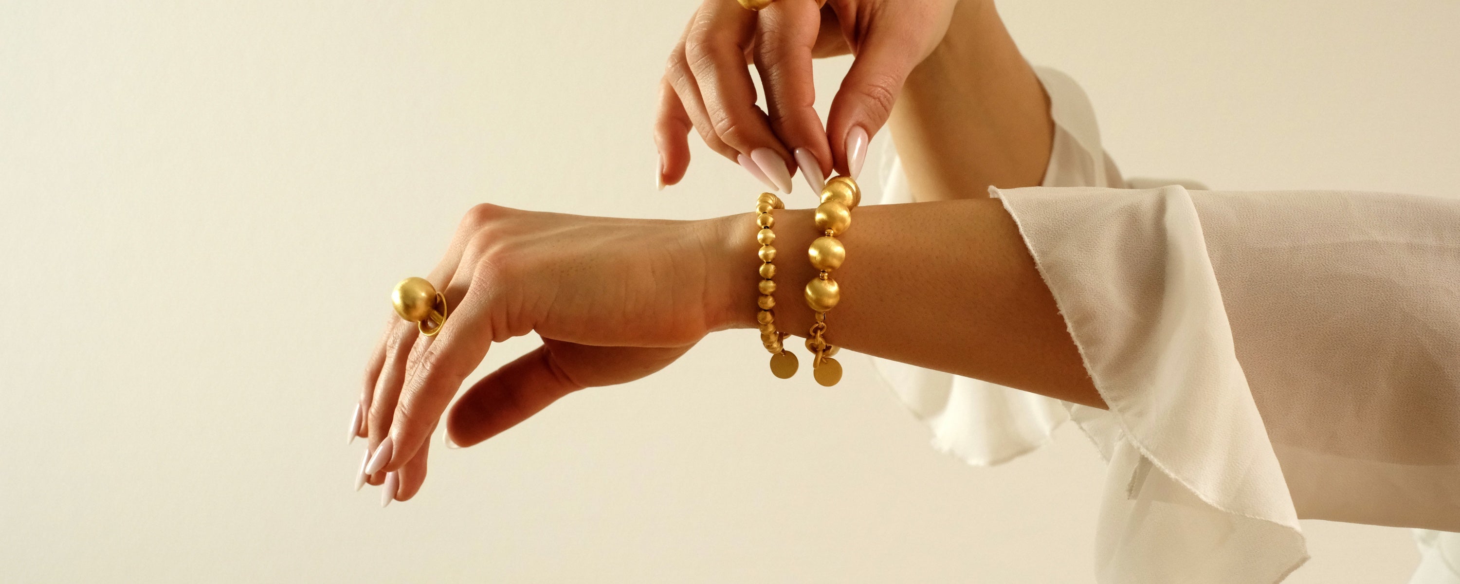 Detailaufnahme von Händen, die elegante goldene Armbänder und einen auffälligen Goldring von Cosytas tragen, kombiniert mit einer leichten weißen Bluse vor minimalistischem Hintergrund.