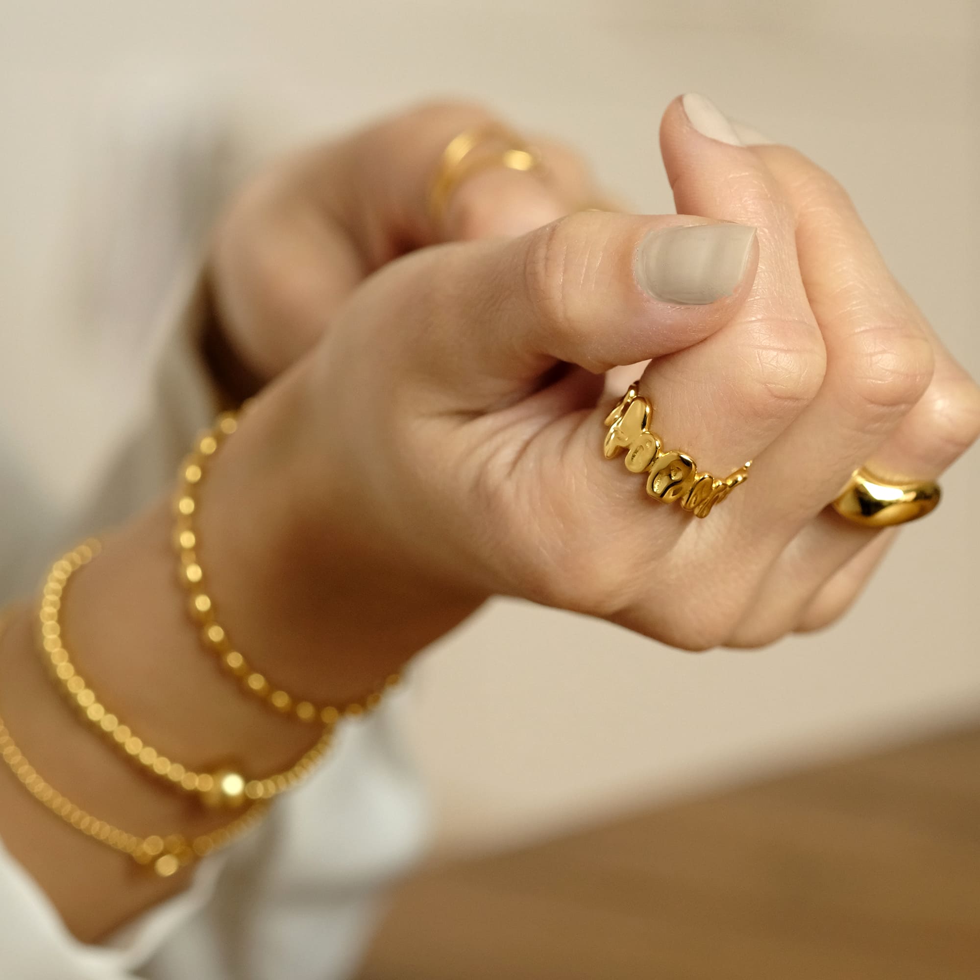 Hände mit goldenen Schmuckstücken, darunter der Cosytas Golden Pebble Ring mit organisch geformten Elementen. Die elegante Kombination mit feinen Armbändern verleiht dem Look eine stilvolle, luxuriöse Note.