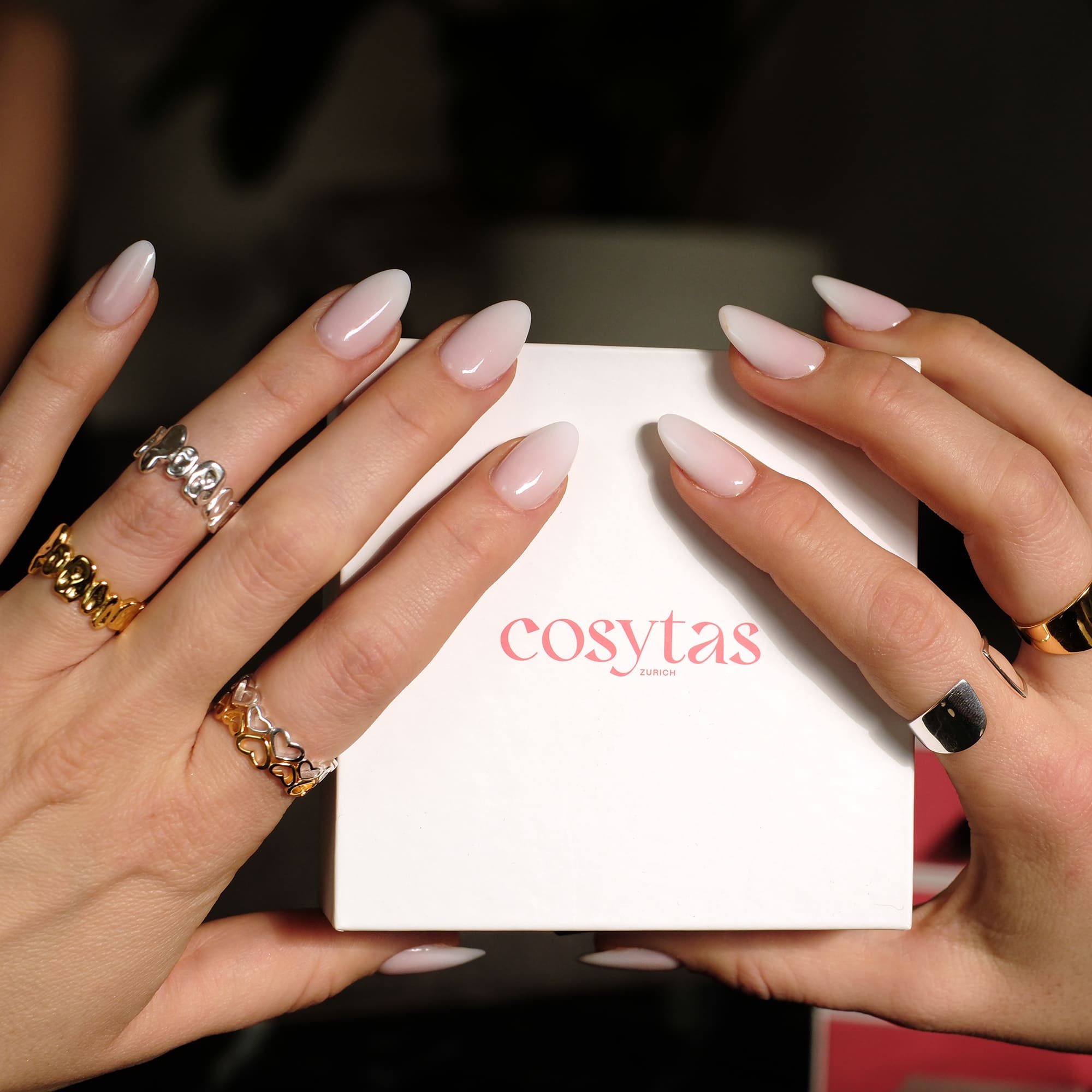 Hände mit perfekt manikürten Nägeln halten eine Cosytas-Schmuckbox. Die Finger sind mit goldenen und silbernen Ringen geschmückt, darunter der Cosytas Heartbeat Ring, der mit seinem filigranen Herzdesign moderne Eleganz verkörpert.
